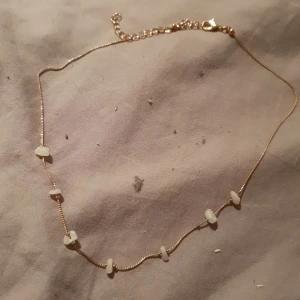 Guldhalsband med vita stenar - Snyggt och tunt guldhalsband med små vita stenar utspridda längs kedjan. Kedjan har en enkel och stilren design med justerbar längd och karbinlås. Perfekt för att ge din outfit en extra touch av elegans.