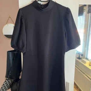 Svart klänning från JDY - Stilren svart klänning från JDY med puffiga korta ärmar och hög krage med knappar i nacken. Klänningen har en rak passform och är tillverkad i ett mjukt, lätt material. Klänningen går ner till knärna ungefär. Klänningen är endast använd fåtal gånger. 