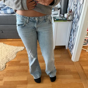 Ljusblå jeans med utsvängda ben - Säljer ett par ljusblå Ginatricot jeans med låg midja och utsvängda ben. Klassisk femficksmodell med snygga detaljer på bakfickorna och knappstängning. Perfekta för en chill och avslappnad stil. Passar dig som gillar vintagevibbar och tidlös denim. Ena av knapparna har trillat av på backfickan och byxorna är lite slitna längst ner annars är de i bra skick💕Nypris 500kr💕