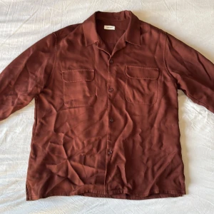 Brun skjorta från Sandro i XL - Snygg brun skjorta från Sandro i storlek XL. Skjortan har klassisk krage, två bröstfickor med lock och knäppning framtill. Långärmad och i ett mjukt, slätt material som känns skönt mot huden. Perfekt för dig som gillar stilren och enkel design. Skrynklig då den legat i garderoben under längre tid men med en tvätt så ska allt vara bra!