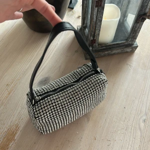Glittrig silver axelväska med strass - Säljer en liten axelväska i silver med massor av glittrande strass över hela väskan. Den har svart axelrem i syntetmaterial och stängs med dragkedja upptill. Perfekt för dig som vill sticka ut och addera extra bling till din outfit. Innerficka finns! Helt ny 
