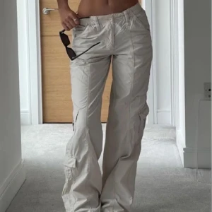 Beiga cargopants Urban Outfitters - Säljer ett par beiga cargopants med flera fickor och dragkedjedetaljer. Byxorna har låg midja och en loose passform som ger en avslappnad vibe. Perfekta för dig som gillar streetstyle och vill ha något bekvämt men ändå trendigt. Lite slitningar längst ner men annars bra skick!! OBS inte min bild 