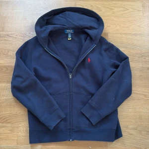 Marinblå zip hoodie från Polo Ralph Lauren - Säljer en marinblå hoodie från Polo Ralph Lauren med dragkedja och huva. Tröjan har en röd broderad logga på bröstet, två fickor framtill och är tillverkad i mjuk bomull. Perfekt för chill dagar och enkel att matcha med det mesta. Den är perfekt nu till hösten och vintern. Original pris 1200. det är en liten fläck vid sidan som man inte ser så mycket när man har den på. om ni inte är nöjda med priset föreslå ett pris och skriv till mig om ni har några frågor
