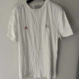 Vit t-shirt från Axel Arigato - Vit t-shirt från Axel Arigato med röd broderad bokstav A och liten broderad fågel på bröstet. Klassisk passform, rund hals och korta ärmar. Storlek XS