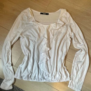 Vit volangblus från Bik Bok - Supersöt vit blus från Bik Bok med långa ärmar och volangdetalj längs ringningen och framtill. Toppens material känns mjukt och lätt, perfekt för en fräsch och stilren look. Passar dig som gillar romantiska detaljer och enkel design.
