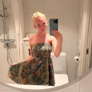 Färgglad axelbandslös kortklänning - Superfin axelbandslös kortklänning med färgstarkt paisleymönster i blått, rött, gult och grönt. Smockad överdel och utsvängd kjol ger en skön och luftig känsla. Perfekt för varma dagar och sommarhäng!