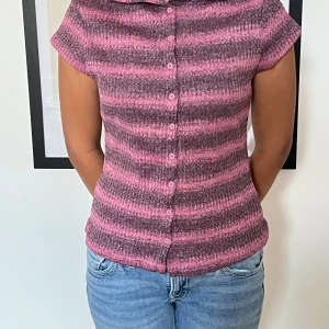 Rosa och lila randig blus med knappar - Supersöt blus med rosa och lila ränder, korta ärmar och knappar hela vägen framtill. Den har en liten krage och är figurnära i modellen. Perfekt för dig som gillar färg och vill sticka ut lite extra.