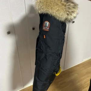 Parajumpers svart parkajacka päls - Svart parkajacka från Parajumpers med stor fluffig päls runt huvan och snygga detaljer som gul rem och patch på ärmen. Jackan har flera fickor, dragkedja och knappar. Perfekt för kalla dagar när du vill hålla stilen och värmen.