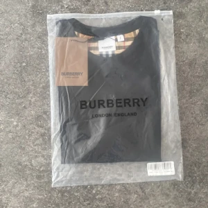 Burberry svart t-shirt med rutiga ärmar - Svart t-shirt från Burberry med klassiska rutiga ärmar i beige, svart, vitt och rött. T-shirten har rund halsringning och är tillverkad i mjuk bomull. Ikonisk och stilren design med Burberry-tag kvar. Perfekt för dig som vill sticka ut med en lyxig touch.