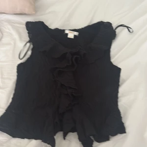 Svart volangblus från Gina Tricot - Supersöt svart blus från Gina Tricot med volangdetaljer framtill och vågig struktur i tyget. Ärmlös modell med lätt rynkade kanter och knytband vid axlarna. Perfekt för dig som gillar en romantisk och trendig look.