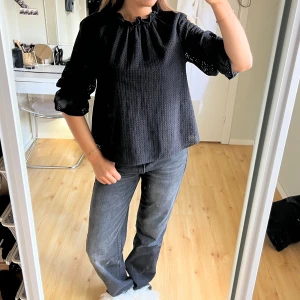 Svart blus med volangärm och brodyr - Säljer en svart blus med broderat hålmönster och volangdetaljer vid ärmsluten. Blusen har trekvartsärm och en söt rynkad halsringning med knytband. Perfekt för dig som gillar en stilren men ändå unik look.