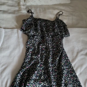 Blommig sommarklänning från Zoul - Supersöt kort blommig klänning från Zoul med tunna axelband och volangdetaljer upptill. Svart botten med små blommor i vitt, rosa, blått och gult. Perfekt för varma dagar och ger en fräsch vibe. Lätt och luftig modell som sitter snyggt.