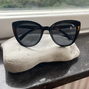 Svarta cat eye-solglasögon Gina Tricot - Snygga svarta cat eye-solglasögon från Gina Tricot med glansig finish och mörka glas. Ramen är markerad och ger en cool retrovibe. Perfekta för att lyfta vilken outfit som helst och skydda ögonen med UV400-filter.