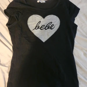 Svart topp med hjärta från Bebe - Svart kortärmad topp från Bebe med ett stort silvrigt hjärta och Bebe-logga framtill. Slimmad passform och rund halsringning. Perfekt för dig som gillar statement-plagg med lite extra attityd. Passar storlek XS-S. sitter pytte lite tajt på mig med S :)