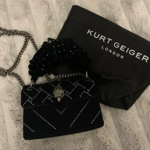 Kurt Geiger väska - Säljer denna så fina väska från Kurt Geiger som är helt oanvänd.  Den är i sammet med silver stenar på. Inga stenar har ramlat av och inga andra defekter, ser ut precis som ny!  Det är ett långt kedjeband så att man kan ha väskan över axeln, sen finns det ett mindre handtag i sammet som man även kan ta av. Nypris ca 4000, och tror inte den går att få tag på längre. Dustbag tillkommer!