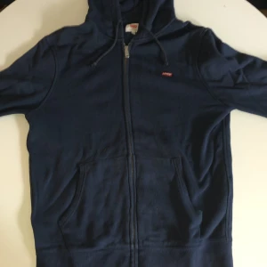 Mörkblå hoodie från Levi's med dragkedja - Säljer en mörkblå hoodie från Levi's i storlek S. Tröjan har hel dragkedja, huva med snörning och två stora fickor framtill. Perfekt för lager-på-lager och chill dagar. Klassisk Levi's-logga på bröstet och mjukt material. 