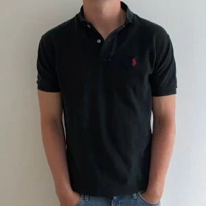 Ralph Lauren Piké - Klassisk svart pikétröja från Polo Ralph Lauren med röd broderad logga på bröstet. Tröjan har korta ärmar, krage och knäppning med knappar. Tillverkad i mjuk bomull som är skön mot huden. Perfekt för en clean och stilren look.