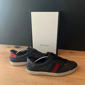 Gucci sneakers med röd och blå detalj - Snygga svarta Gucci sneakers i skinn med klassiska röda och blå ränder på sidorna. Skorna har snörning, rund tå och mönstrad sula med Gucci-logga. Baktill har ena skon röd och den andra blå detalj i ormskinnsmönster. Perfekta för dig som gillar lyxig streetstyle.