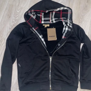 Svart Burberry hoodie med dragkedja - Svart hoodie från Burberry med klassiskt rutigt foder i huvan och broderad logga på bröstet. Tröjan har dragkedja framtill, två fickor och snörning i huvan. Den är i ny skick och prislapp finns kvar. Snabb affär pris diskuteras 