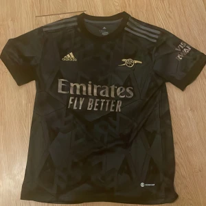 Svart Arsenal fotbollströja Adidas - Snygg svart Arsenal fotbollströja från Adidas med guldiga detaljer och tryck. Tröjan har korta ärmar, diskret geometriskt mönster och klassiska Adidas-ränder på axlarna. Materialet är lätt och andas, perfekt för träning eller match.