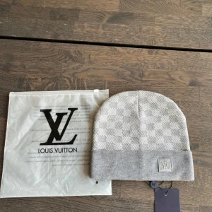 Grå Louis Vuitton rutig mössa - Exklusiv grå mössa från Louis Vuitton med klassiskt rutigt mönster och vikt kant. LV-logga framtill i broderad patch. Tillverkad i mjukt material som känns lyxigt och stilrent. Perfekt accessoar för att lyfta din vinterlook.