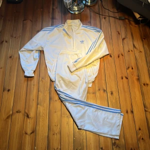 Vit Adidas tracksuit med blå ränder - Snygg vit Adidas vindjacka med klassiska blå ränder längs ärmarna och dragkedja framtill. Jackan har hög krage, ribbade muddar och två fickor med dragkedja. Perfekt för en sportig och retro vibe. Adidas logga broderad på bröstet. Med ett par feta addidas byxor som gör detta till en tracksuit. Storleken är M och den är oversized så den är mer som en L båda jackan och byxor kommer med skriv för frågor och pruta för en snabb affär 