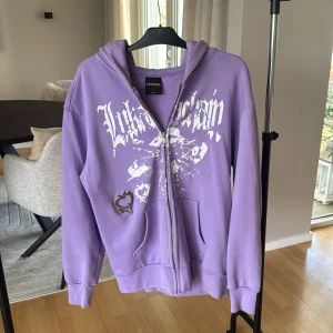 Lila hoodie från LUKAMACHAIN med tryck - Lila hoodie från LUKAMACHAIN med vit grafisk text och splattertryck framtill. Dragkedja hela vägen, huva och känguruficka. Snygg metallisk hjärt-detalj på ena sidan. Oversized fit och mjukt material, perfekt för chill dagar.
