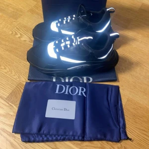 Svarta sneakers från Dior med reflex - Snygga svarta sneakers från Dior med reflekterande detaljer och vit snörning. Skorna har en chunky sula och meshpartier för extra komfort. Perfekta för dig som vill sticka ut med exklusiv stil och coola detaljer.