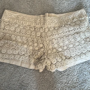 Beige spetsiga shorts med broderi - Superfina beige shorts med detaljerad spets och broderade blommor över hela tyget. De har normal passform, knapp och dragkedja framtill samt fickor. Perfekta för sommarens varma dagar när du vill ha en romantisk och chill vibe.