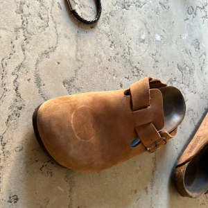 Bruna Birkenstock Boston i mocka - Säljer ett par klassiska Birkenstock Boston i brun mocka med justerbart spänne i metall. Skorna har rund tå och platt sula i gummi för extra komfort. Perfekta för dig som gillar en avslappnad och stilren look.