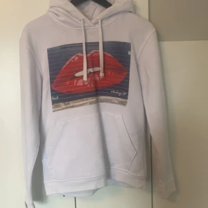 Vit hoodie med röd läppar-print - Vit hoodie från The Park Support med stor grafisk print av röda läppar på bröstet. Klassisk känguruficka, huva med snörning och långa ärmar. Mjuk och skön bomullsblandning, perfekt för chill dagar💞