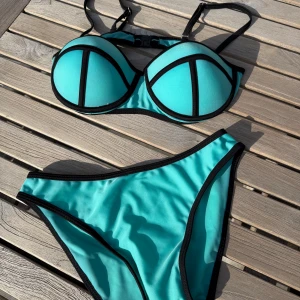 Turkos bikini från MANYA med svarta kanter - Snygg turkos bikini från MANYA med svarta kontrastkanter och vadderad bygel-bh. Klassisk bikiniunderdel med hög midja och bekväm passform. Perfekt för strandhäng och poolparty. Justerbara axelband och spänne i ryggen.