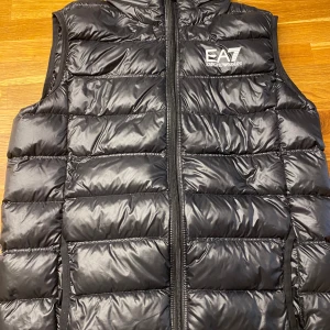 Svart dunväst från Emporio Armani EA7 - Säljer en svart, quiltad dunväst från Emporio Armani EA7. Västen har en stilren design med logga på bröstet och dragkedja framtill. Perfekt att slänga över en hoodie för en clean look. Materialet är glansigt och känns lätt men ändå värmande. Luva finns i dragkedjan vid nacken om man vill ha det. Pris kan diskuteras via snabbaffär.