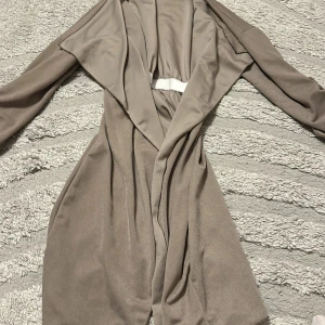 Beige trenchcoat med draperad krage - Snygg beige trenchcoat med draperad, öppen krage och långa ärmar. Jackan har en avslappnad passform och är tillverkad i ett mjukt, lätt material som ger ett stilrent intryck. Perfekt för lager-på-lager och en clean look.