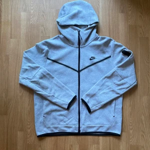 Nike Tech Fleece i färgen grå - Nike Tech Fleece hoodiejacka i ljusgrått med svart logga på bröstet och hel dragkedja. Jackan har huva, svarta detaljer längs dragkedjan och ärmslut samt en praktisk ficka med dragkedja på ena ärmen. Perfekt för chill eller träning. Köptes för 1300kr