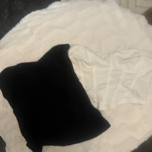 Svart bandeau & vit korsett topp - Säljer två snygga toppar! En svart bandeau i ribbat tyg, perfekt för lager-på-lager eller som den är. Den andra är en vit korsett-topp med markerade sömmar och bystparti, ger en riktigt cool siluett. Båda är axelbandslösa och passar till många olika looks.