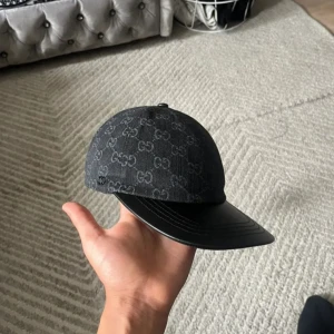 Svart Gucci keps med GG-mönster - Snygg svart keps från Gucci med klassiskt GG-monogram över hela ovandelen. Skärmen är i svart skinn och kepsen har justerbar rem baktill. Materialet är en mix av bomull och skinn, perfekt för dig som vill sticka ut med en lyxig accessoar.