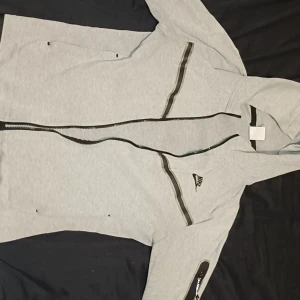 Grå nike tech hoodie  - Snygg grå hoodie från Nike med svarta detaljer och dragkedjor. Hoodien har huva, flera fickor med dragkedja och Nike-logga på bröstet. Perfekt för en sportig och avslappnad stil.