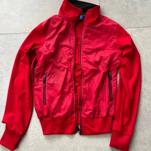 Röd vindjacka från Adidas, stl S - Snygg röd vindjacka från Adidas med hög krage och svarta dragkedjor på fickorna. Jackan har ribbade muddar och nederkant, samt hel dragkedja framtill. Perfekt för dig som gillar sportig och clean stil.