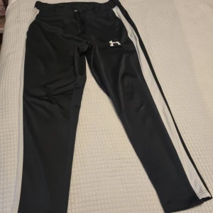 Svarta träningsbyxor Under Armour - Svarta träningsbyxor från Under Armour i storlek XL. Byxorna är vita längs sidorna. De har snörning i midjan och logga på låret. Se bilder för mått. Se gärna mina andra annonser och skapa bundles. Inga byten.