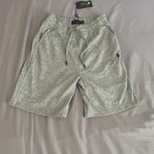 Grå shorts från Polo Ralph Lauren - Snygga grå shorts från Polo Ralph Lauren med elastisk midja och snörning. Klassisk logga broderad på ena benet. Mjukt material i bomull, perfekt för chill dagar eller träning. Fickor på sidorna och avslappnad passform.