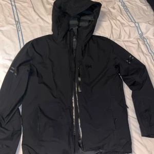 Svart pufferjacka från Helly Hansen - Svart pufferjacka från Helly Hansen med huva och dragkedja framtill. Jackan har quiltad insida, två sidofickor med dragkedja och diskreta reflexdetaljer på ärmarna. Perfekt för kalla dagar och riktigt snygg streetstil.