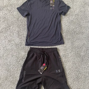 Under armour set - Svart kortärmad t-shirt från Under Armour med diskret logga på bröstet och tryckt text längs sidan. Tillverkad i lätt och ventilerande polyester, perfekt för träning. Snygg, sportig look med rund hals och bekväm passform. Den är i helt nytt och den är aldrig amvänt.