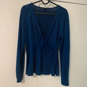 Blå omlottopp med volangdetalj - Supersnygg blå omlottopp med långa ärmar och volang längs den djupa v-ringningen. Knyt i sidan för en extra touch. Perfekt för dig som gillar en feminin och trendig stil. Storlek M.