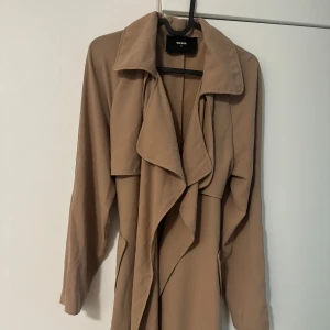 Beige trenchcoat från Bik Bok - Snygg beige trenchcoat från Bik Bok med långa ärmar, bred krage och knytband i midjan. Jackan har två fickor och ett avslappnat, flowy fall. Perfekt för dig som vill ha en stilren och trendig look till vardags. Storlek M.