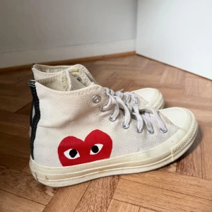 Converse x CDG Play vita high sneakers - Ikoniska Converse Chuck Taylor All Star high sneakers i samarbete med Comme des Garçons Play. Vita canvas med klassisk röd hjärtdetalj på sidan, snörning och rund tå. Snygg kontrast med svart rand baktill och gummisula. Perfekta för dig som gillar streetstyle. Skorna är ganska använda och smutsiga men jag kan absolut tvätta dom innan dom skickas!! Skorna kommer med lådan. Kan absolut skicka mer bilder på skorna om ni är intresserade i dom