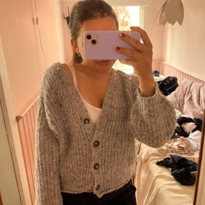 Grå stickad kofta med knappar - Mysig grå stickad kofta med stora svarta knappar och bred ribbad struktur. Koftan är croppad och har långa ärmar med lite oversized passform. Storleken är 152 men passar som en xs eftersom den är stretchig och lite oversized!