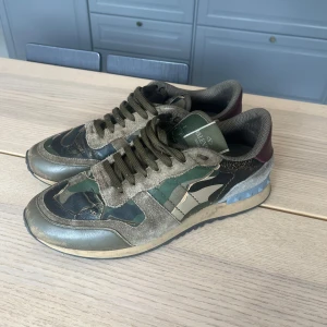 Valentino Rockrunners - Snygga Valentino Rockrunner sneakers med camouflage-mönster i grönt, svart och beige. Skorna har detaljer i mocka, skinn och textil samt vinröda hälkappor och blå nitar på sulan. Klassisk snörning och platt sula. Storlek 42. Tveka inte med frågor!