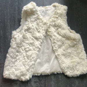 Vit pälsväst från H&M, stl 140 - Fluffig och mjuk vit pälsväst från H&M i storlek 140. Västen har ett stilrent snitt utan knäppning och är helfodrad med slät insida. 