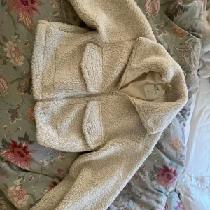 Beige teddyjacka från Outfish, S - Supermjuk och croppad teddyjacka i beige från Outfish. Jackan har stora fickor framtill, dragkedja och bred krage. Perfekt för dig som gillar en chill och cozy vibe. Passformen är loose och ärmarna är långa för extra mysfaktor.